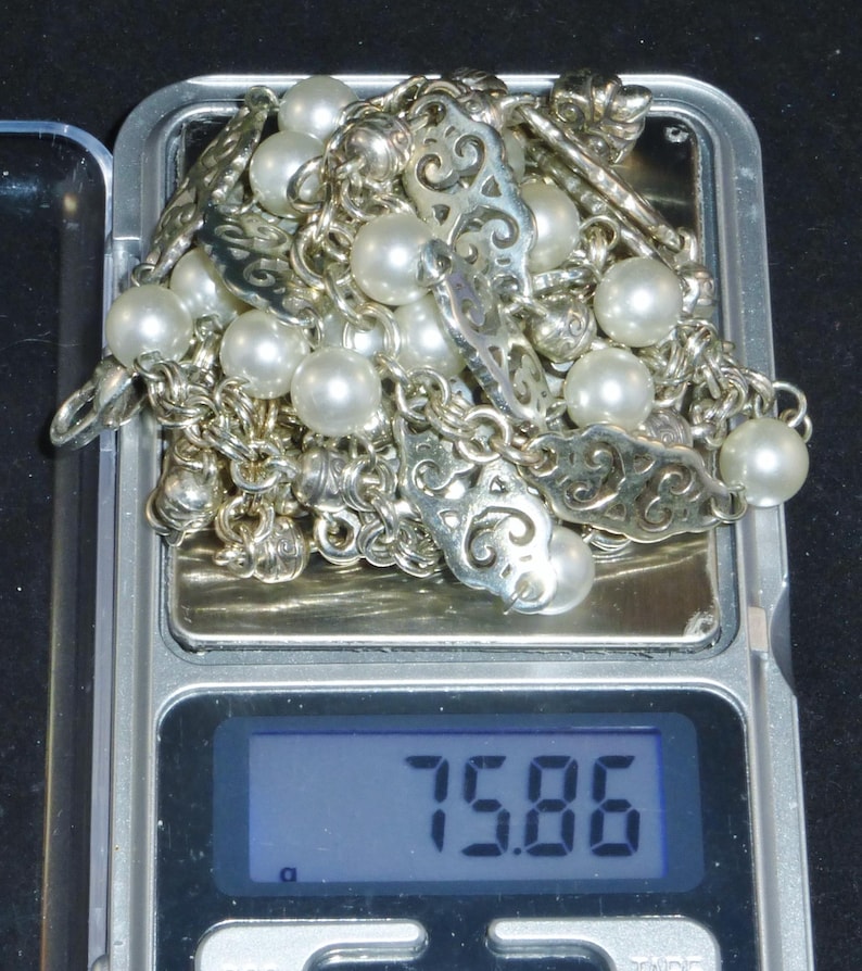 May include: A silver chain necklace with white pearls and ornate silver charms. The necklace is on a digital scale that reads 75.86.
