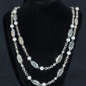 May include: A silver chain necklace with white pearls and a delicate filigree design. The necklace is double-stranded and has a long length.