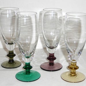 May include: Four clear champagne flutes with colored bases. The bases are in shades of green, purple, and yellow. The stems have a decorative design. The glasses are empty and sit on a white surface.