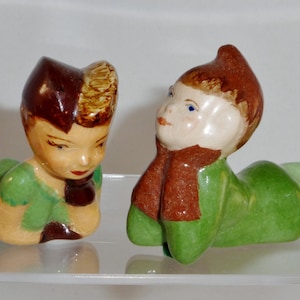 May include: Two ceramic figurines of children dressed in green and brown clothing. The figurines are lying on their stomachs with their heads turned to the side.