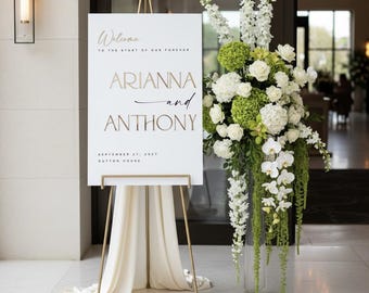 May include: A white wedding welcome sign on a gold metal easel with cream fabric draped underneath. The sign reads: Welcome to the start of our forever, Arianna and Anthony, September 27, 2027, Dutton House. Beside it is a tall glass vase with white flowers.