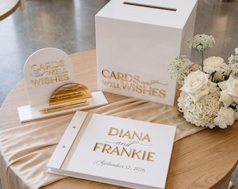 May include: A white square card box with a slot, a matching guest book labeled Diana and Frankie September 12, 2026, and an arched sign with gold lettering reading Cards and Well Wishes, accompanied by gold pens and a white floral arrangement.