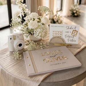 May include: A white instant camera, a beige guest book with gold lettering reading Louise and Oliver October 6, 2027, and a sign that reads Shake it like a Polaroid Picture, displayed with a white floral arrangement and two gold pens.