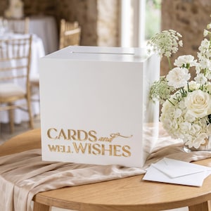 Könnte beinhalten: Eine quadratische weiße Box mit einem Schlitz an der Oberseite zum Sammeln von Umschlägen. Auf der Vorderseite befindet sich der goldfarbene Schriftzug "CARDS and WELL WISHES" in einer Mischung aus Serifenschrift und Schreibschrift.