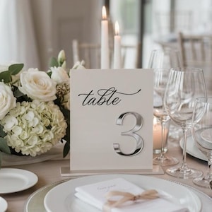 Op de afbeelding: Een rechthoekig wit tafelnummerbordje met het woord table in zwarte cursieve letters en een groot zilveren cijfer 3. Het bordje staat op een gedekte tafel met witte borden, een servet met een lint, glaswerk en witte bloemen.