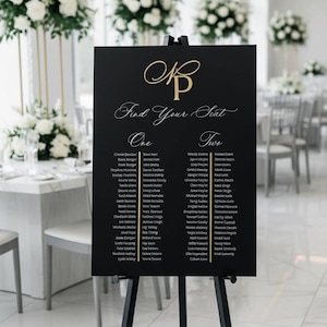May include: A black rectangular wedding seating chart sign on a black tripod easel. The sign features gold lettering with the monogram KP at the top, the text Find Your Seat, and two columns labelled One and Two listing guest names in gold.