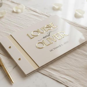 May include: A cream-colored guest book with gold embossed lettering that reads "LOUISE and OLIVER" and "OCTOBER 6, 2027". The book features a side binding with two gold metal rivets and is accompanied by a matching gold pen resting beside it.