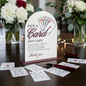 May include: An arched white sign with a maroon base featuring a graphic of playing cards and the text: PICK A Card ANY CARD PLEASE WRITE A MESSAGE AND SIGN YOUR NAME SO WE CAN REMEMBER THIS DAY WHENEVER WE PLAY Thank you. Includes two pens.