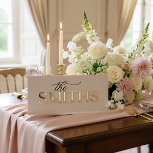 May include: A wedding reception table setting with a floral arrangement, candles, and a sign that reads "the SMITHS" in gold lettering. The table is covered with a pink runner and set with gold flatware.
