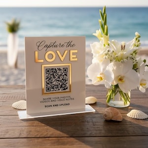 Könnte beinhalten: Ein weißes rechteckiges Schild mit goldener Schrift, die Capture the LOVE liest, mit einem zentralen QR-Code und dem Text SHARE YOUR PHOTOS, VIDEOS AND VOICE NOTES, SCAN AND UPLOAD, neben einer Glasvase mit weißen Blumen.