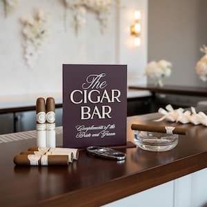 Könnte beinhalten: Ein dunkelviolettes rechteckiges Schild mit weißer Aufschrift The CIGAR BAR Complements of the Bride and Groom. Das Schild wird zusammen mit mehreren braunen Zigarren, einem silbernen Zigarrenschneider und einem klaren Glasaschenbecher präsentiert.