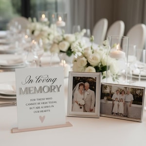 Op de afbeelding: Een wit rechthoekig bord met zwarte en zilveren tekst In loving MEMORY FOR THOSE WHO CANNOT BE HERE TODAY, BUT ARE ALWAYS IN OUR HEARTS met een klein roze hartje, tentoongesteld naast twee zilveren fotolijstjes op een tafel.