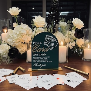 May include: A dark green arched sign with gold text reading PICK A Card ANY CARD PLEASE WRITE A MESSAGE AND SIGN YOUR NAME SO WE CAN REMEMBER THIS DAY WHENEVER WE PLAY Thank you, featuring a graphic of playing cards, resting on a gold base.
