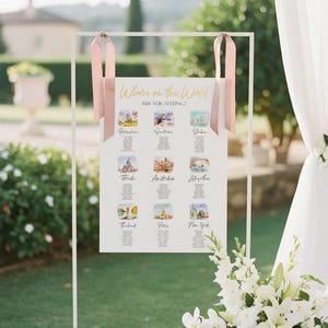 May include: A white rectangular wedding seating chart sign suspended from a thin white metal frame by two dusty pink ribbons. The sign features the gold text Where in the World ARE YOU SITTING? above nine watercolor illustrations of global cities.