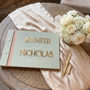 May include: A sage green guest book with a textured tan spine and gold metal accents. The cover features the raised gold text JENNIFER and NICHOLAS with the date AUGUST 25, 2027. A gold pen rests beside the book, which is placed on a beige fabric runner.