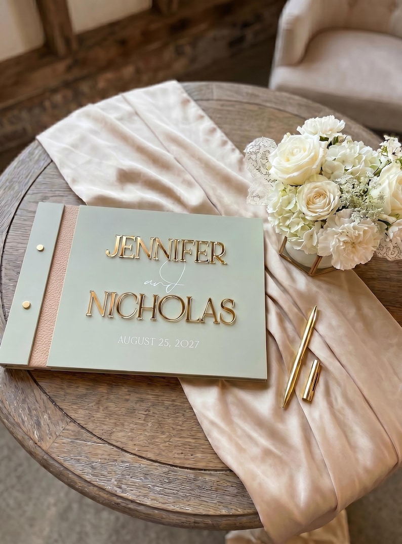May include: A sage green guest book with a textured tan spine and gold metal accents. The cover features the raised gold text JENNIFER and NICHOLAS with the date AUGUST 25, 2027. A gold pen rests beside the book, which is placed on a beige fabric runner.