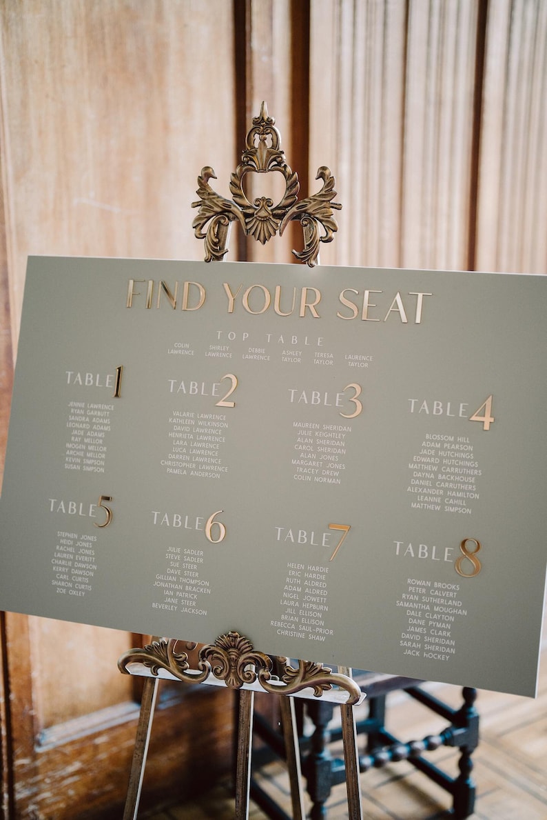 May include: A sage green seating chart with gold lettering and accents. The sign reads "FIND YOUR SEAT" and lists names under table numbers 1 through 8. The sign is displayed on a decorative gold easel.