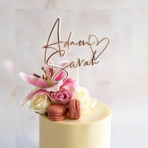 Könnte beinhalten: Ein weißer und roségoldener Tortenaufleger mit den Namen Adam und Sarah in Schreibschrift und einem Herzumriss. Der Aufleger befindet sich auf einer Torte, die mit rosa Lilien, weißen und rosa Rosen sowie zwei rosa Macarons verziert ist.