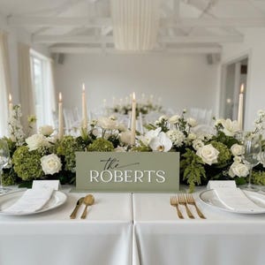 May include: A sage green rectangular table sign with the white text The ROBERTS, placed on a white tablecloth. The sign is surrounded by white roses, green hydrangeas, and tall taper candles, with gold cutlery and white plates on the table.