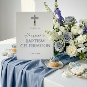 May include: A baptism celebration sign with a silver cross and text that reads "Welcome to Harrison's Baptism Celebration June 16, 2027." The sign is on a table with a blue tablecloth, cupcakes, and cookies. A floral arrangement is in the background.