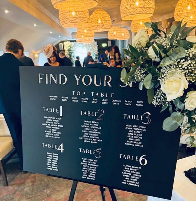 May include: A black wedding seating chart with white lettering, reading "Find Your Seat." The chart lists names under table numbers 1-6. The sign is on a black easel, with floral arrangements and hanging lights in the background.