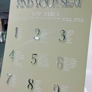 May include: A wedding seating chart with the words "Please Find Your Seat" in silver lettering. The chart lists names under table numbers 1 through 9. The background is a light beige color, and the overall design is elegant.