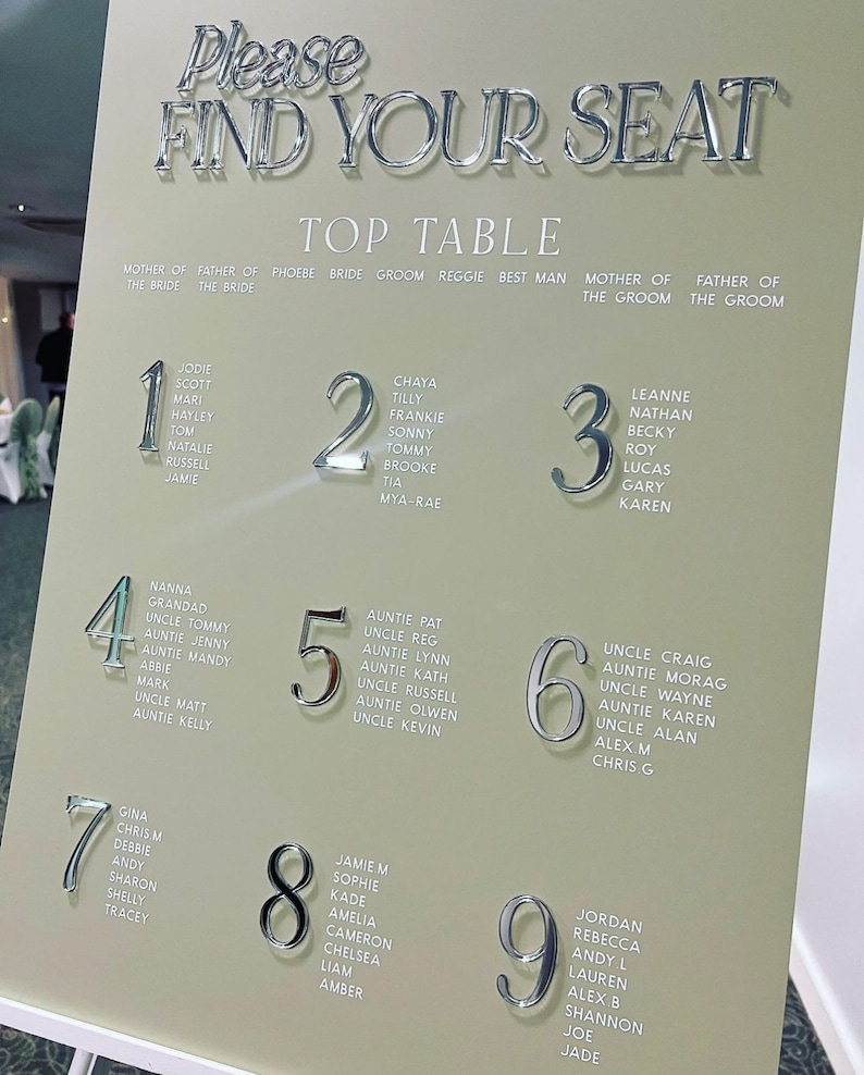 May include: A wedding seating chart with the words "Please Find Your Seat" in silver lettering. The chart lists names under table numbers 1 through 9. The background is a light beige color, and the overall design is elegant.