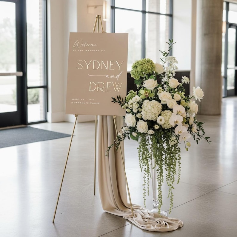 May include: A wedding welcome sign on a gold easel with the names "SYDNEY and DREW". The sign is beige with gold lettering. A floral arrangement of white and green flowers is next to the sign, with a beige fabric draped over the easel.
