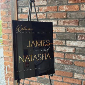May include: A black acrylic wedding welcome sign with gold lettering. The sign reads "Welcome to our wedding celebration" at the top, with the names "JAMES and NATASHA" in large gold letters. The date "25th October 2025" is at the bottom.