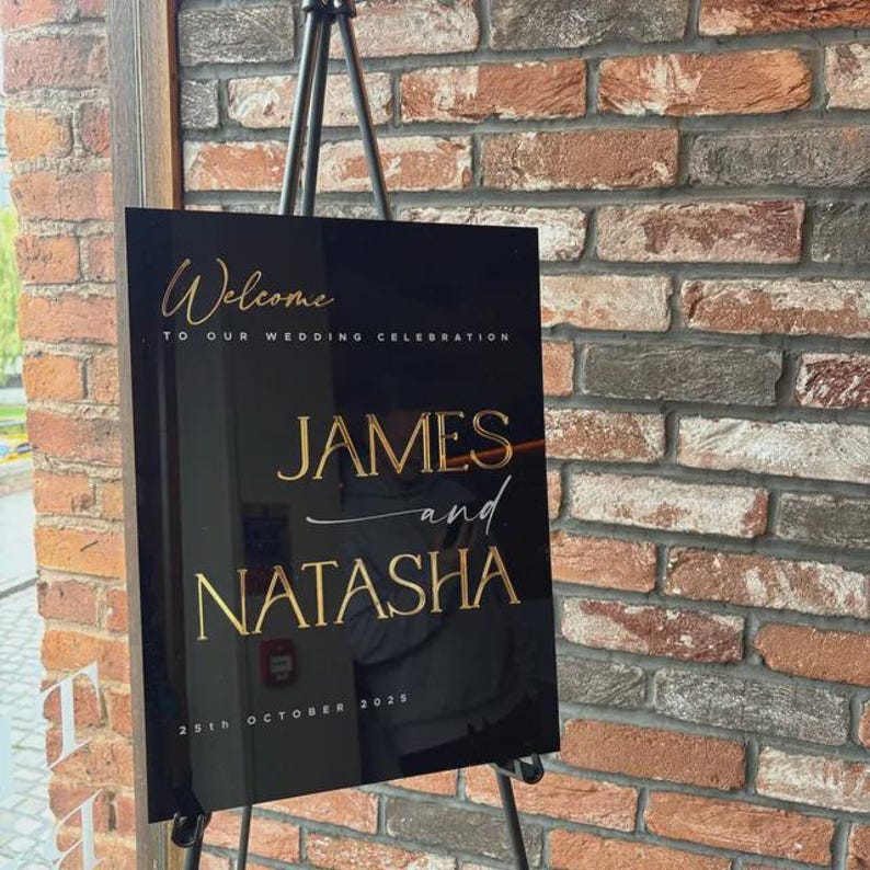 May include: A black acrylic wedding welcome sign with gold lettering. The sign reads "Welcome to our wedding celebration" at the top, with the names "JAMES and NATASHA" in large gold letters. The date "25th October 2025" is at the bottom.
