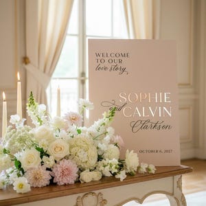 May include: A wedding reception sign with the text "WELCOME TO OUR love story" and the names "SOPHIE and CALVIN Clarkson" with the date "OCTOBER 6, 2027". The sign is on a table with a floral arrangement and candles.