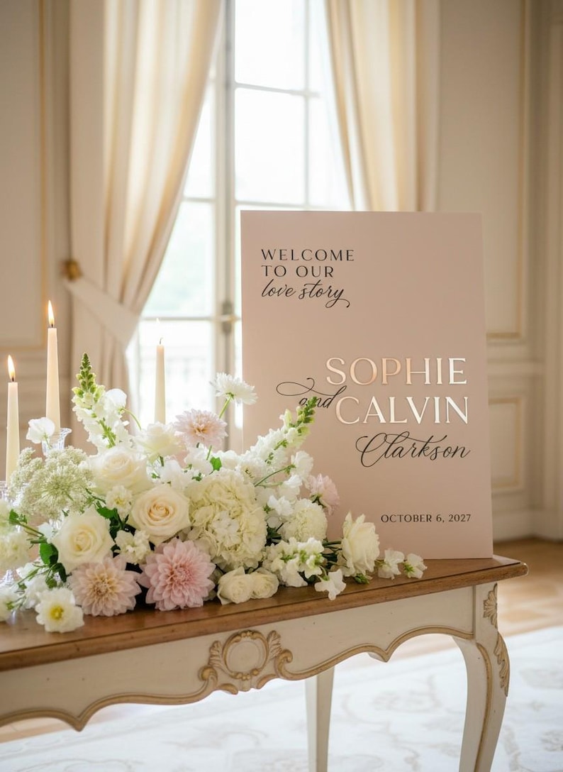 May include: A wedding reception sign with the text "WELCOME TO OUR love story" and the names "SOPHIE and CALVIN Clarkson" with the date "OCTOBER 6, 2027". The sign is on a table with a floral arrangement and candles.