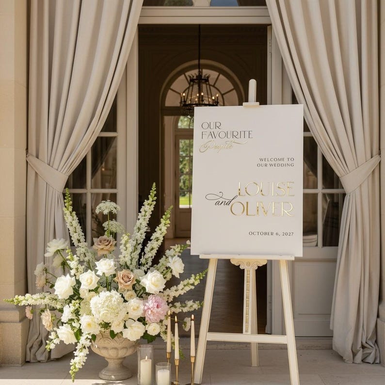 May include: A wedding welcome sign on a white easel with gold lettering. The sign reads "Our Favourite People, Welcome to Our Wedding, Louise and Oliver, October 6, 2027." A floral arrangement and candles are in front of the sign.