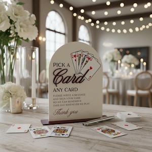May include: An arched off-white sign with a dark brown base featuring a graphic of playing cards and the text: PICK A Card ANY CARD, PLEASE WRITE A MESSAGE AND SIGN YOUR NAME SO WE CAN REMEMBER THIS DAY WHENEVER WE PLAY, Thank you.