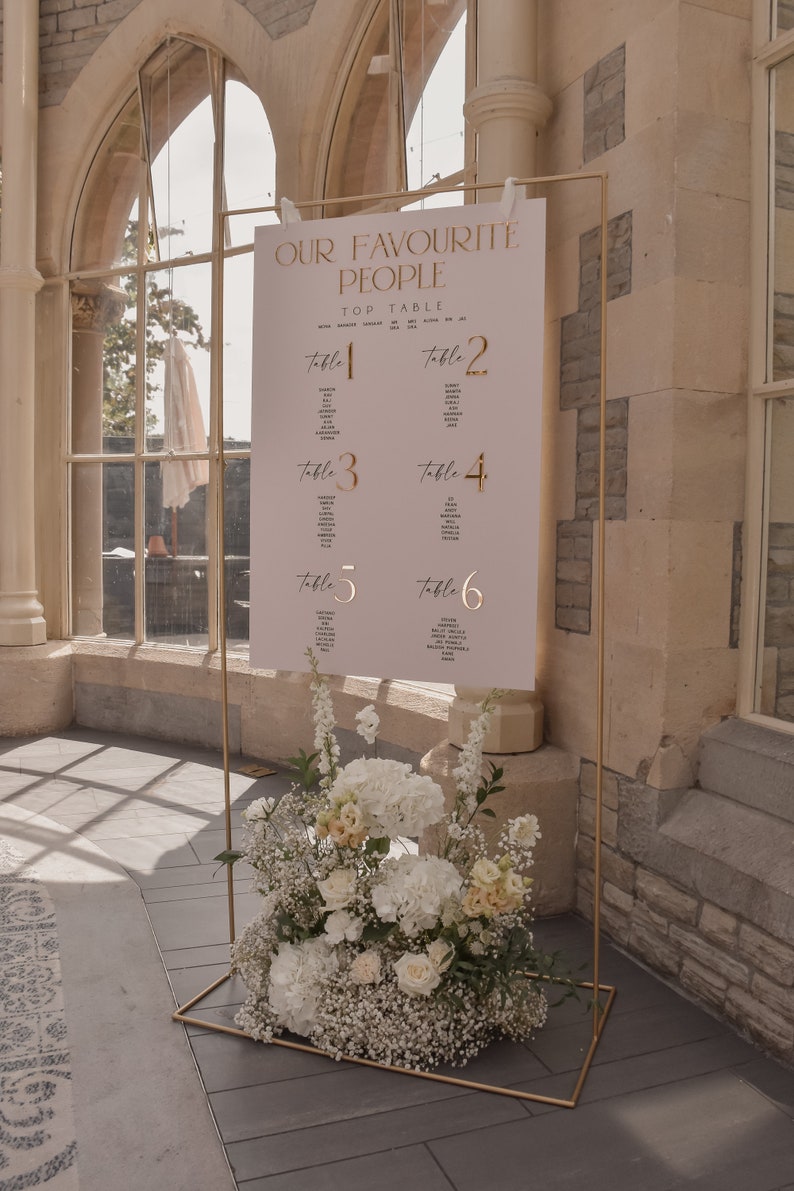 May include: A gold metal frame holds a sign with the text "OUR FAVOURITE PEOPLE" and table assignments. The sign is decorated with gold lettering. A floral arrangement of white and cream flowers sits at the base of the frame. The setting is a light-filled room.