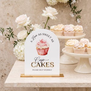 May include: A white arched sign with a gold base featuring a watercolor cupcake illustration and black text that reads A Love As Sweet As Cupcakes PLEASE HELP YOURSELF. The sign is displayed alongside white cupcakes and white roses.