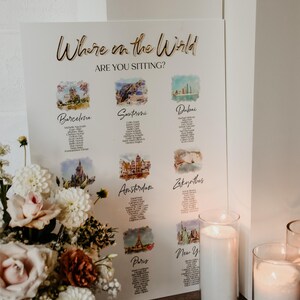 May include: A white rectangular wedding seating chart sign with the gold text Where in the World ARE YOU SITTING? featuring watercolor illustrations of global cities like Barcelona, Santorini, Dubai, Amsterdam, Zakynthos, and Paris with guest lists.