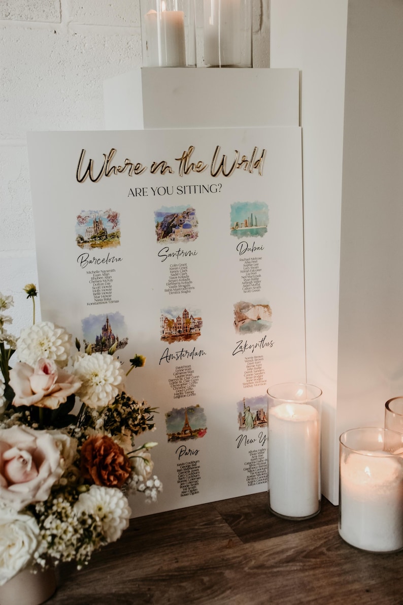 May include: A white rectangular wedding seating chart sign with the gold text Where in the World ARE YOU SITTING? featuring watercolor illustrations of global cities like Barcelona, Santorini, Dubai, Amsterdam, Zakynthos, and Paris with guest lists.