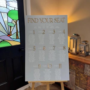 May include: A light gray seating chart with gold lettering that reads "FIND YOUR SEAT". The chart lists names under table numbers 1 through 11. The chart is displayed on a wooden easel.