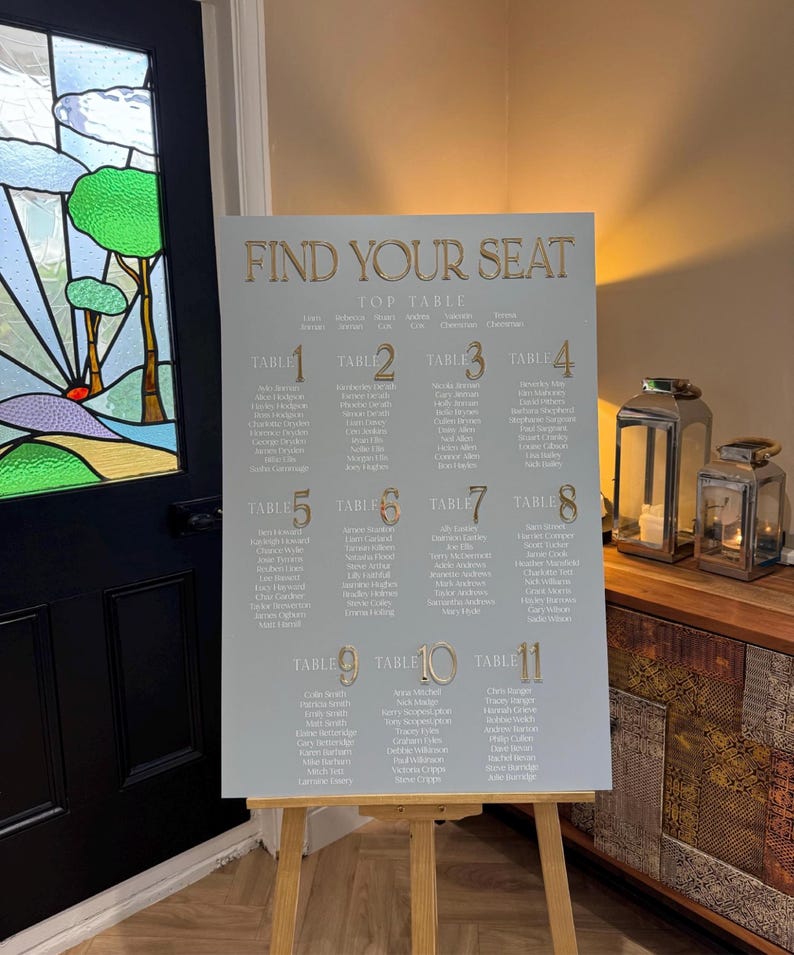 May include: A light gray seating chart with gold lettering that reads "FIND YOUR SEAT". The chart lists names under table numbers 1 through 11. The chart is displayed on a wooden easel.
