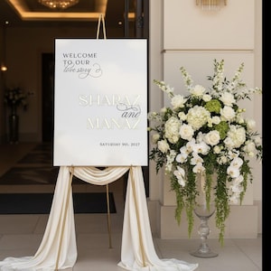 May include: A white wedding welcome sign on a gold easel with draped fabric. The sign reads "Welcome to our love story" and features the names "Sharaz and Manaz" with the date "Saturday 9th, 2027". A large floral arrangement of white flowers is to the right.