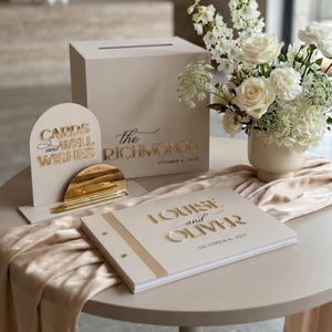 Op de afbeelding: Een beige trouwkaartenbox met de tekst "the RICHMONDS" en "OCTOBER 6, 2027", een boogvormig bord met "CARDS and WELL WISHES", een gouden pennenset en een gastenboek "LOUISE and OLIVER" met de datum "OCTOBER 6, 2027".