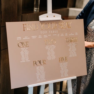May include: A blush pink wedding seating chart with gold lettering. The sign reads "FIND YOUR SEAT" at the top, followed by table numbers and guest names. The sign is displayed on a white easel.