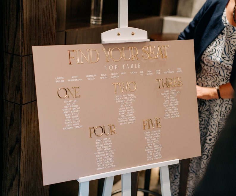 May include: A blush pink wedding seating chart with gold lettering. The sign reads "FIND YOUR SEAT" at the top, followed by table numbers and guest names. The sign is displayed on a white easel.