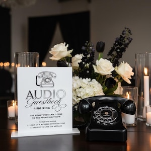 Puede incluir: Un tel&eacute;fono rotatorio negro vintage se encuentra sobre una superficie oscura junto a un letrero que dice "Audio Guestbook". El letrero es blanco con texto negro y un gr&aacute;fico de tel&eacute;fono. Un arreglo floral de rosas blancas, tulipanes negros y vegetaci&oacute;n est&aacute; en el fondo.