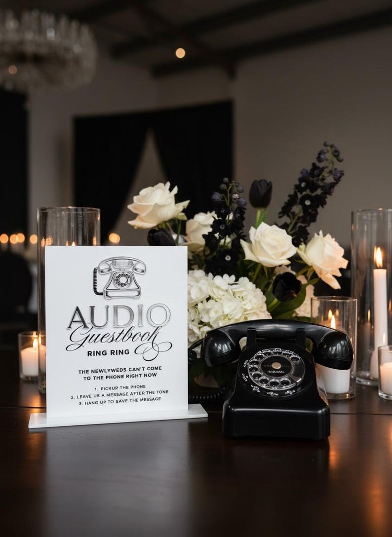 Puede incluir: Un tel&eacute;fono rotatorio negro vintage se encuentra sobre una superficie oscura junto a un letrero que dice "Audio Guestbook". El letrero es blanco con texto negro y un gr&aacute;fico de tel&eacute;fono. Un arreglo floral de rosas blancas, tulipanes negros y vegetaci&oacute;n est&aacute; en el fondo.