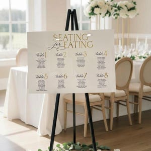 May include: A rectangular white acrylic wedding seating chart sign with gold lettering that reads SEATING for EATING, listing guests for tables one through eight. The sign is displayed on a black tripod easel stand on a wooden floor.