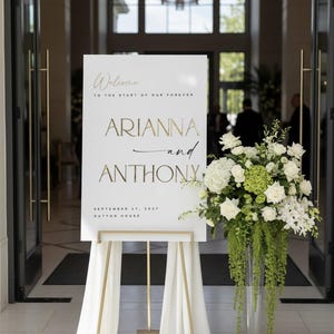 May include: A white welcome sign with gold lettering, announcing "Arianna and Anthony" and the date "September 27, 2027". The sign is on a gold easel, draped with white fabric, and next to a tall vase of white and green flowers.
