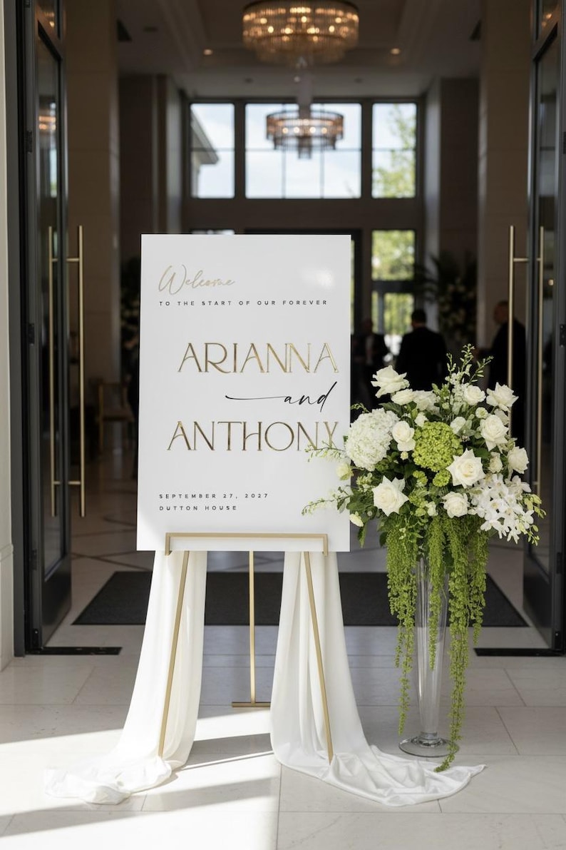 May include: A white welcome sign with gold lettering, announcing "Arianna and Anthony" and the date "September 27, 2027". The sign is on a gold easel, draped with white fabric, and next to a tall vase of white and green flowers.