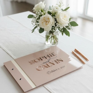May include: A blush pink guest book with a glossy finish featuring the embossed text "SOPHIE and CALVIN" and "October 6, 2027" in a metallic rose gold font. The book has a side binding with two silver metal rivets and two rose gold pens.